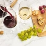 Top down view of a cheese board with a variety of cheeses, crackers and fruit along with a few glasses of red and white wine.