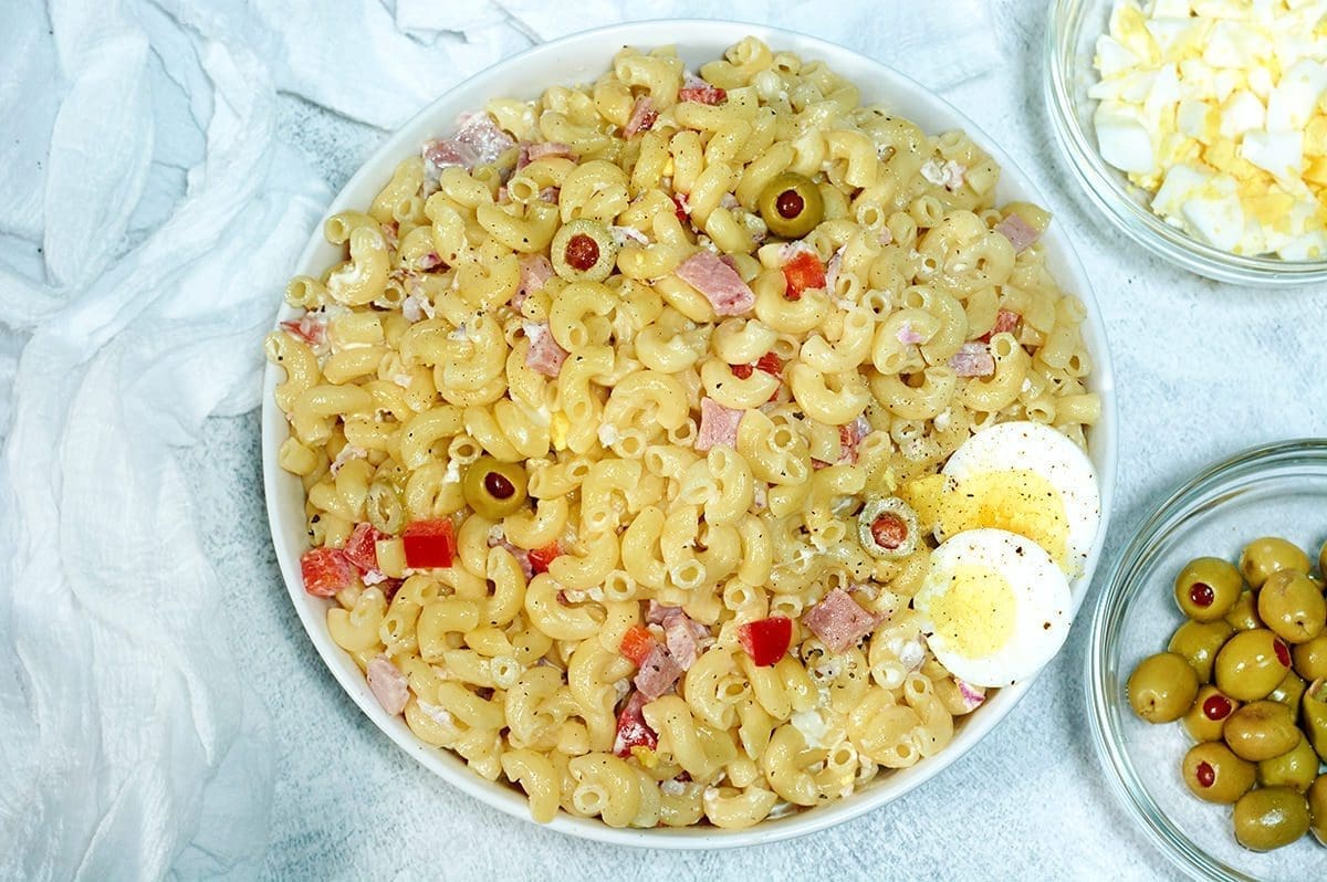 Ensalada de Coditos (Puerto Rican Macaroni Salad) in a large white bowl surrounded by small bowls of hard boiled eggs, cilantro and olives.