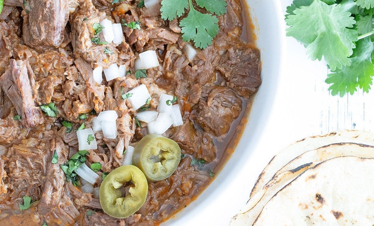 Close up of Birria in a white bowl topping with onions, pickled jalapenos and cilantro.