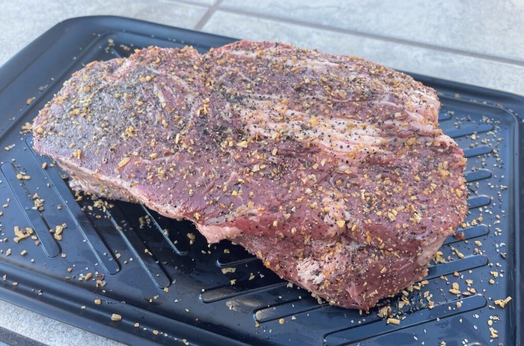 Seasoned and uncooked chuck roast on a black plastic plate.